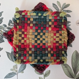 May include: A square, woven hot pad in a patchwork design. The pad features a mix of red, green, brown, and yellow threads. The edges are a darker red, and the corners are slightly raised. The background is a floral print.