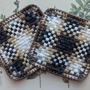 May include: Two square woven potholders. The potholders feature a woven pattern in shades of white, tan, brown, and navy blue. The edges are finished with a brown border. The potholders are displayed on a botanical print background.