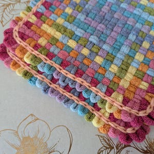 May include: Close-up of three colorful woven coasters. The coasters are made of woven yarn in shades of pink, orange, yellow, green, blue, and purple. Each coaster has a scalloped edge in a darker pink color and a light pink border.
