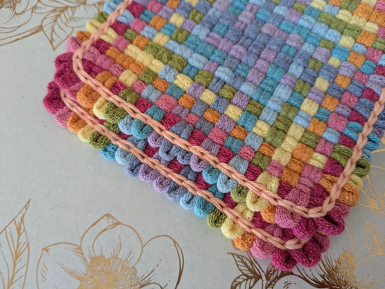 May include: Close-up of three colorful woven coasters. The coasters are made of woven yarn in shades of pink, orange, yellow, green, blue, and purple. Each coaster has a scalloped edge in a darker pink color and a light pink border.