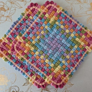 May include: Three colorful, woven square potholders. Each potholder features a woven pattern in shades of pink, orange, yellow, green, blue, and purple. The edges are trimmed with a pink scalloped border. The potholders are stacked on top of each other.