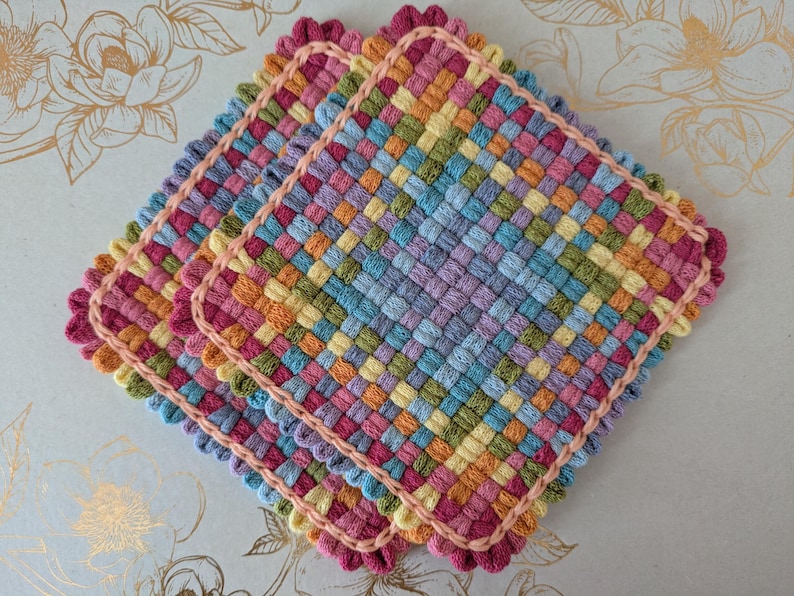May include: Three colorful, woven square potholders. Each potholder features a woven pattern in shades of pink, orange, yellow, green, blue, and purple. The edges are trimmed with a pink scalloped border. The potholders are stacked on top of each other.