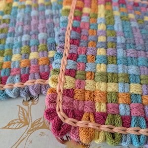 May include: Close-up of a handwoven textile with a colorful, checkered pattern. The fabric features squares of blue, green, yellow, orange, pink, and purple. A peach-colored, looped border frames the edges of the textile.