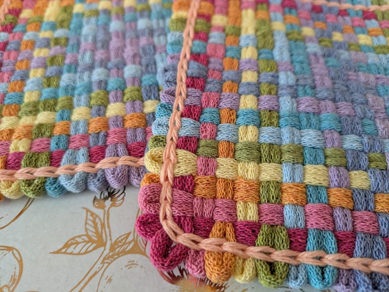 May include: Close-up of a handwoven textile with a colorful, checkered pattern. The fabric features squares of blue, green, yellow, orange, pink, and purple. A peach-colored, looped border frames the edges of the textile.