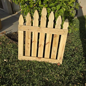 May include: A small, wooden picket fence with pointed tops. The fence is made of light-colored wood and is set on a bed of green grass. The fence is designed to be decorative and is likely used for a garden or yard.