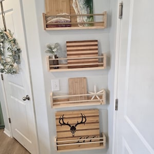 May include: Four wooden shelves with a metal rack on each shelf, mounted on a white wall. The shelves are holding various cutting boards, including a wooden cutting board with a deer skull design, a striped cutting board, a wooden cutting board with a white and green resin design, and a plain wooden cutting board.