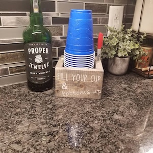 May include: A wooden cup holder with the text "Fill your cup & bottoms up" on the front. A stack of blue plastic cups is in the holder. A bottle of Proper Twelve Irish Whiskey is on the counter.