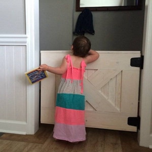May include: A young child in a pink, grey, turquoise, and purple striped dress stands in front of a white wooden gate with a white X-shaped design. The child is holding a book in their left hand.