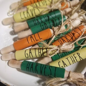 May include: A collection of small, decorative rolling pins in a white bowl. Each pin is painted in fall colors like orange, green, and yellow, with words like "Leaves" and "Harvest" written on them. They are tied with twine.
