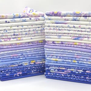 May include: Two tall stacks of folded fabric squares arranged in a gradient from light to dark. The top pieces feature floral patterns in shades of purple, blue, and white, while the bottom pieces transition into solid light blue and deep royal blue.