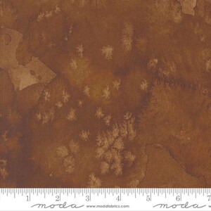 May include: A brown fabric with a mottled, speckled pattern. The fabric has a subtle, faded look, with lighter brown spots scattered across the surface. The fabric is textured and has a slightly rough feel.