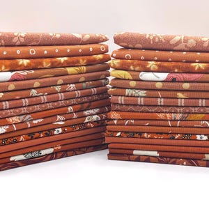 May include: Two stacks of folded fabric in shades of orange and brown. The fabric features various floral and geometric patterns. The stacks are neatly arranged, showcasing the different designs and textures of the material.