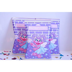 May include: Three rectangular snack bags featuring a purple brick wall background with a light blue and purple star pattern. Each bag displays a cartoon character with purple hair, a light blue dress, and wings, holding a wand with the text AALIYAH'S STREET.