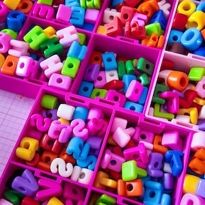 May include: A pink plastic organizer box filled with a large assortment of colorful plastic alphabet letter beads and square spacer beads in various shades of blue, green, yellow, orange, pink, purple, and white, arranged in separate compartments.