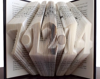 May include: A book sculpture with the word "LOVE" cut into the pages. The pages are folded to create a 3D effect, with the letters standing out. The book has a black cover and is displayed on a dark wooden surface.