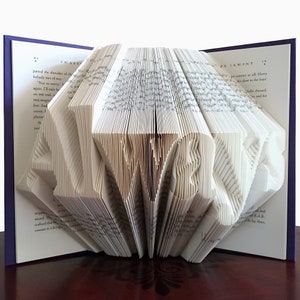 May include: A hardcover book with pages folded to spell out the word "LOVE". The book has a purple cover and the pages are white with black text. The book is open, revealing the folded word in the center.