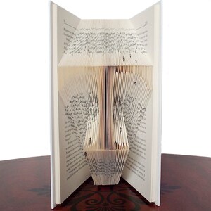 May include: A book art sculpture, also known as a book folding, in the shape of a tree. The book is open and the pages are folded to create the shape of a tree. The book is on a wooden surface.