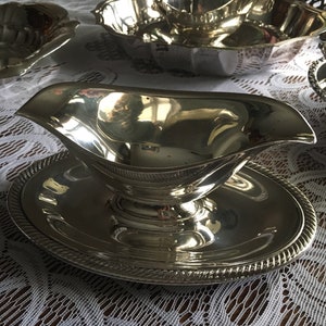 May include: A silver gravy boat with a matching round base, featuring a decorative rope-like border. The boat has a curved spout and a handle. Other silver serving dishes are visible in the background.