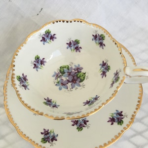 May include: A white teacup and saucer set with a floral violet pattern. The teacup and saucer have a scalloped edge and are trimmed with gold. The teacup is filled with a cluster of violets.