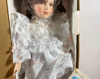 May include: A porcelain doll in a box, wearing a white dress with lace trim and pink accents. The doll has brown hair, a veil, and a tag that reads "Milestone". The doll is a collectible item.