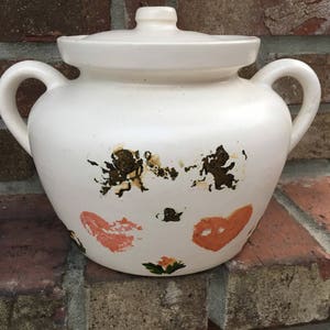 May include: White ceramic jar with a lid and two handles. The jar is decorated with brown and orange abstract designs. The jar is sitting on a brick surface.