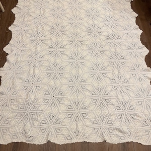 May include: A large, white, crocheted blanket with a geometric starburst pattern. The blanket is rectangular with a scalloped edge. The intricate design features repeating six-pointed star shapes, creating a textured and visually appealing surface.