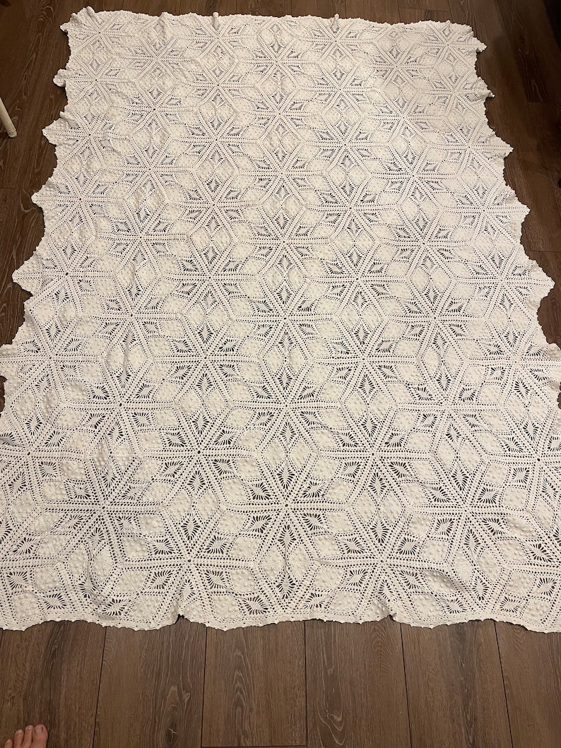 May include: A large, white, crocheted blanket with a geometric starburst pattern. The blanket is rectangular with a scalloped edge. The intricate design features repeating six-pointed star shapes, creating a textured and visually appealing surface.
