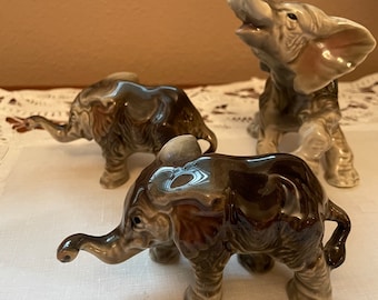 May include: Three ceramic elephant figurines in varying poses. The elephants are primarily shades of gray and brown, with detailed textures. One elephant has its trunk raised, while the others have their trunks lowered.