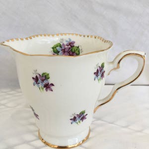 May include: White porcelain creamer with a gold rim and handle, adorned with small purple and blue floral clusters. The creamer has a scalloped edge and a footed base, showcasing a vintage design.