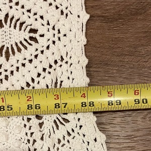 May include: Close-up of a white, handmade crochet doily with a floral design. The doily is measured with a yellow tape measure, showing a width of approximately 84 to 91 inches.