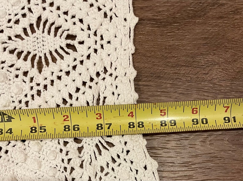 May include: Close-up of a white, handmade crochet doily with a floral design. The doily is measured with a yellow tape measure, showing a width of approximately 84 to 91 inches.