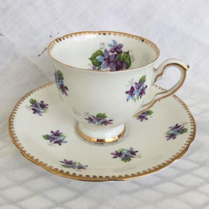 May include: A white teacup and saucer set with a gold trim and a floral violet design. The teacup has a handle and sits on a matching saucer. The set is made of porcelain.
