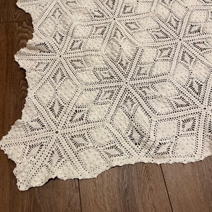 May include: A close-up of a white, crocheted blanket with a geometric starburst pattern. The blanket is made of a soft, textured yarn and has a scalloped edge. The blanket is lying on a dark wooden surface.