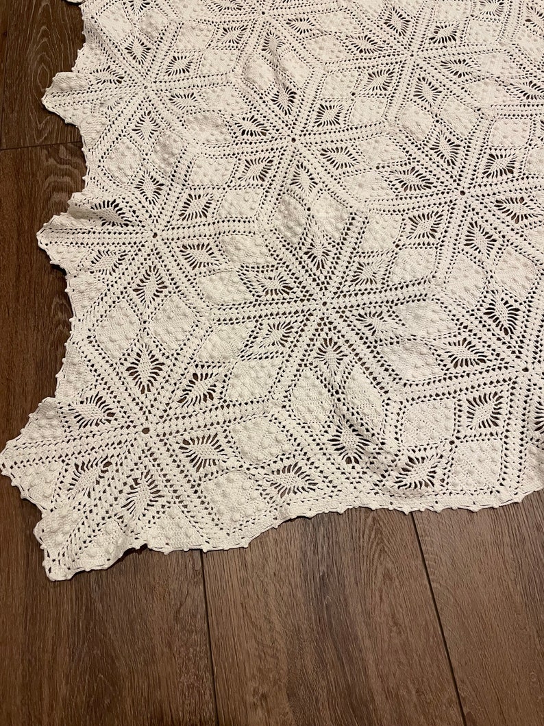 May include: A close-up of a white, crocheted blanket with a geometric starburst pattern. The blanket is made of a soft, textured yarn and has a scalloped edge. The blanket is lying on a dark wooden surface.