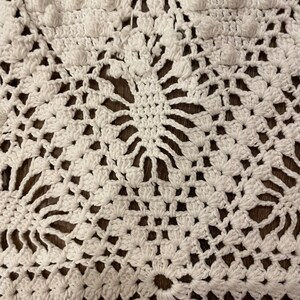 May include: Close-up of a white, handmade crochet doily. The intricate design features floral patterns and raised, textured bobbles. The doily is made of a soft, white yarn and is displayed on a dark brown surface.