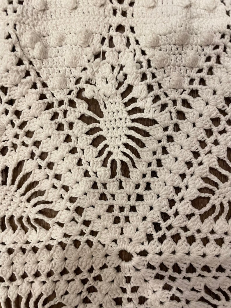 May include: Close-up of a white, handmade crochet doily. The intricate design features floral patterns and raised, textured bobbles. The doily is made of a soft, white yarn and is displayed on a dark brown surface.