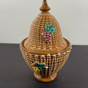 May include: A brown ceramic jar with a woven basket design, featuring a lid and base. The jar is adorned with grape cluster and leaf accents in green, yellow, and purple. The jar has a pedestal base and a pointed finial on the lid.