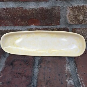 May include: A rectangular, off-white ceramic serving dish with rounded edges. The dish has a glossy finish and a slightly irregular surface, suggesting a handmade quality. The background is a weathered brick wall.