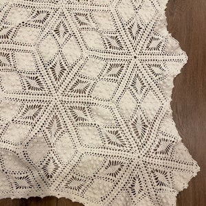May include: A white, handmade crochet blanket with a detailed geometric pattern. The blanket features a repeating design of star-like shapes and diamond patterns, creating a textured surface. The edges are scalloped, adding a decorative finish.