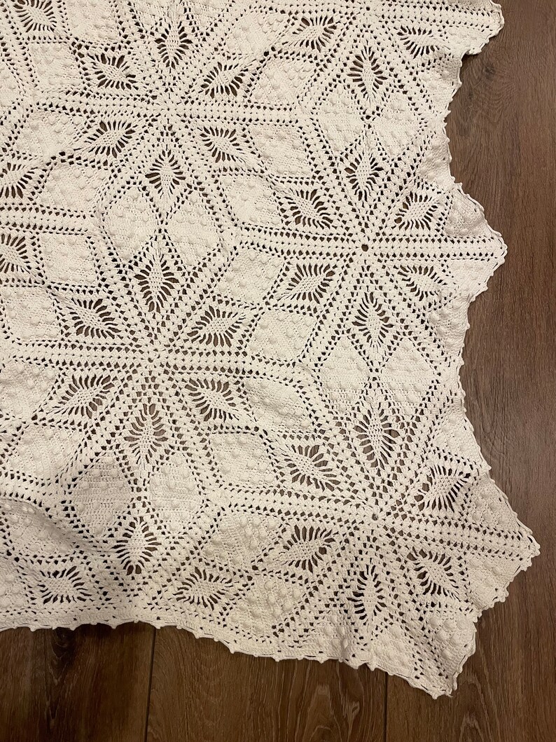 May include: A white, handmade crochet blanket with a detailed geometric pattern. The blanket features a repeating design of star-like shapes and diamond patterns, creating a textured surface. The edges are scalloped, adding a decorative finish.