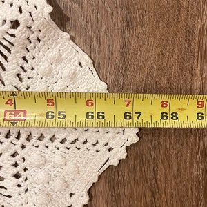 May include: A white, crocheted doily with a scalloped edge, partially visible against a wooden surface. A yellow measuring tape shows a length of approximately 64 to 69 inches.