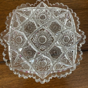 May include: Clear, square-shaped glass dish with intricate geometric patterns and a scalloped edge. The design features starburst and diamond shapes, creating a textured, decorative surface. The dish is likely for serving or display.