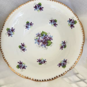 May include: White ceramic plate with a scalloped gold rim and a floral pattern. The plate features clusters of purple and blue flowers with green leaves, evenly spaced around the plate's surface. The center of the plate has a larger floral arrangement.