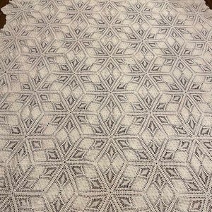 May include: A large, off-white crocheted blanket with a repeating geometric starburst pattern. The blanket is made of a soft, textured yarn and has a delicate, openwork design. The blanket is spread out on a dark surface.