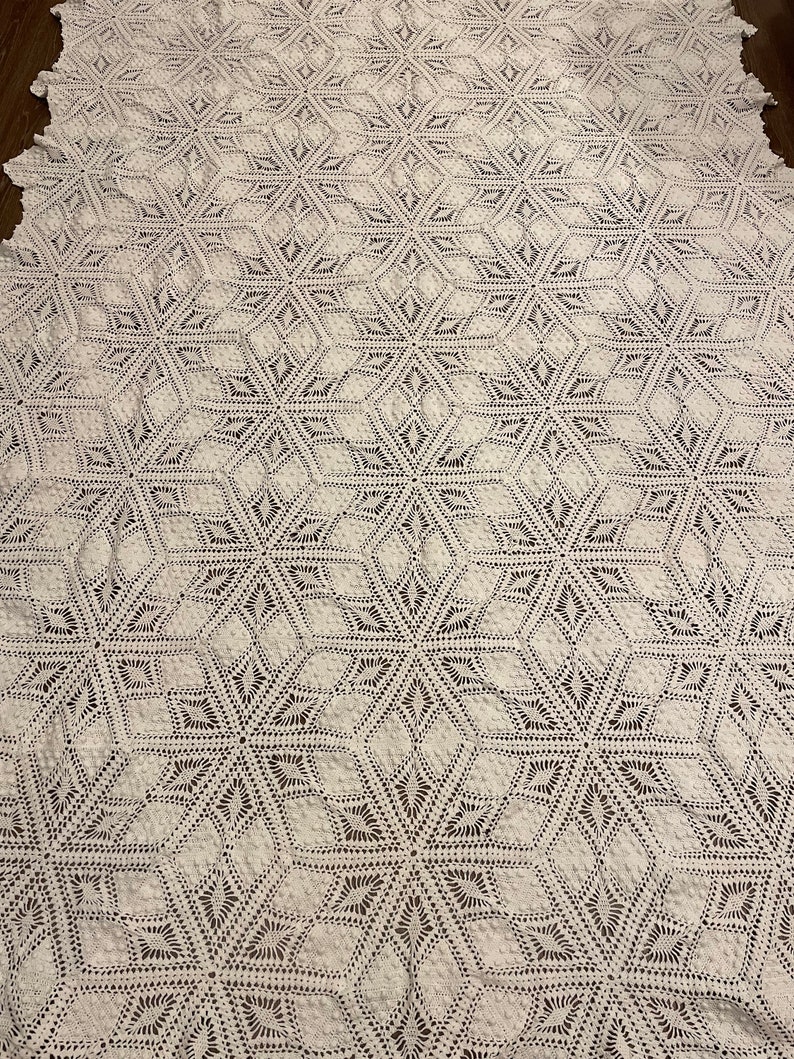May include: A large, off-white crocheted blanket with a repeating geometric starburst pattern. The blanket is made of a soft, textured yarn and has a delicate, openwork design. The blanket is spread out on a dark surface.