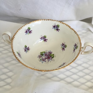 May include: Cream-colored teacup with two handles, adorned with purple and blue floral patterns and a gold rim. The teacup has a scalloped edge and is likely made of porcelain.