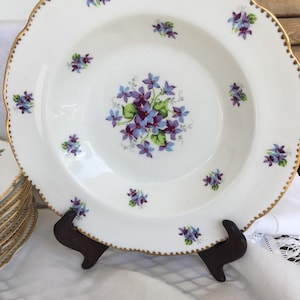 May include: White porcelain plate with a scalloped edge and gold trim. The plate features a central floral design of purple and blue flowers, with smaller floral clusters around the rim. A stack of similar plates is visible.