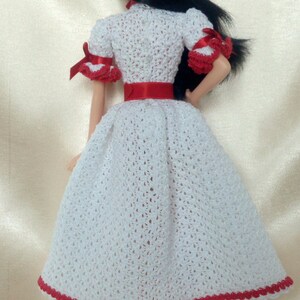May include: A doll wearing a handmade white crochet dress with red accents. The dress features a red ribbon belt and trim, and short sleeves with red bows. The doll has black hair.