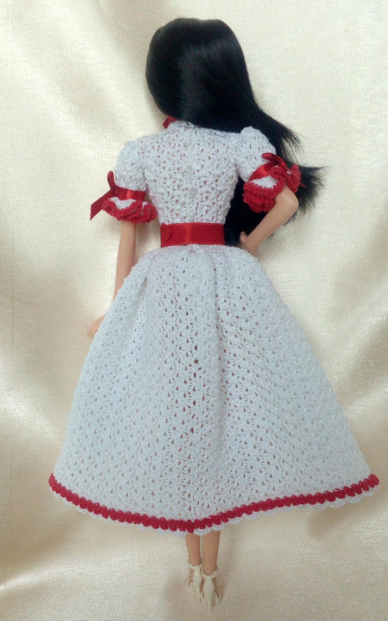 May include: A doll wearing a handmade white crochet dress with red accents. The dress features a red ribbon belt and trim, and short sleeves with red bows. The doll has black hair.