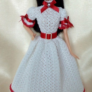 May include: A doll wearing a white crocheted dress with red trim and accents. The dress features a red belt and bow, and the doll has black hair and a gold headband. The dress is a vintage style.